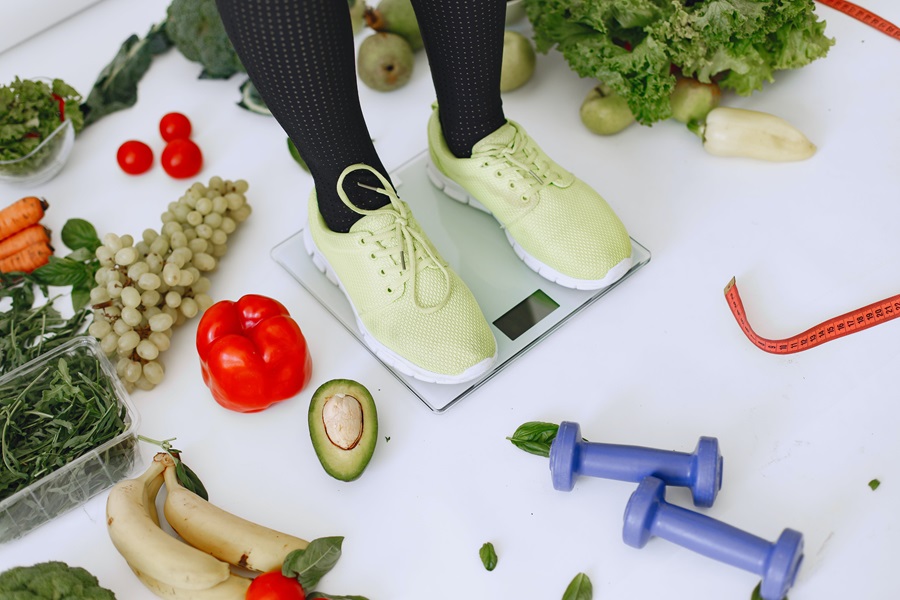 Core De Force Meal Plan Tips Close Up of a Person Standing on a Scale with Veggies Surrounding the Scale and Two Dumbbells